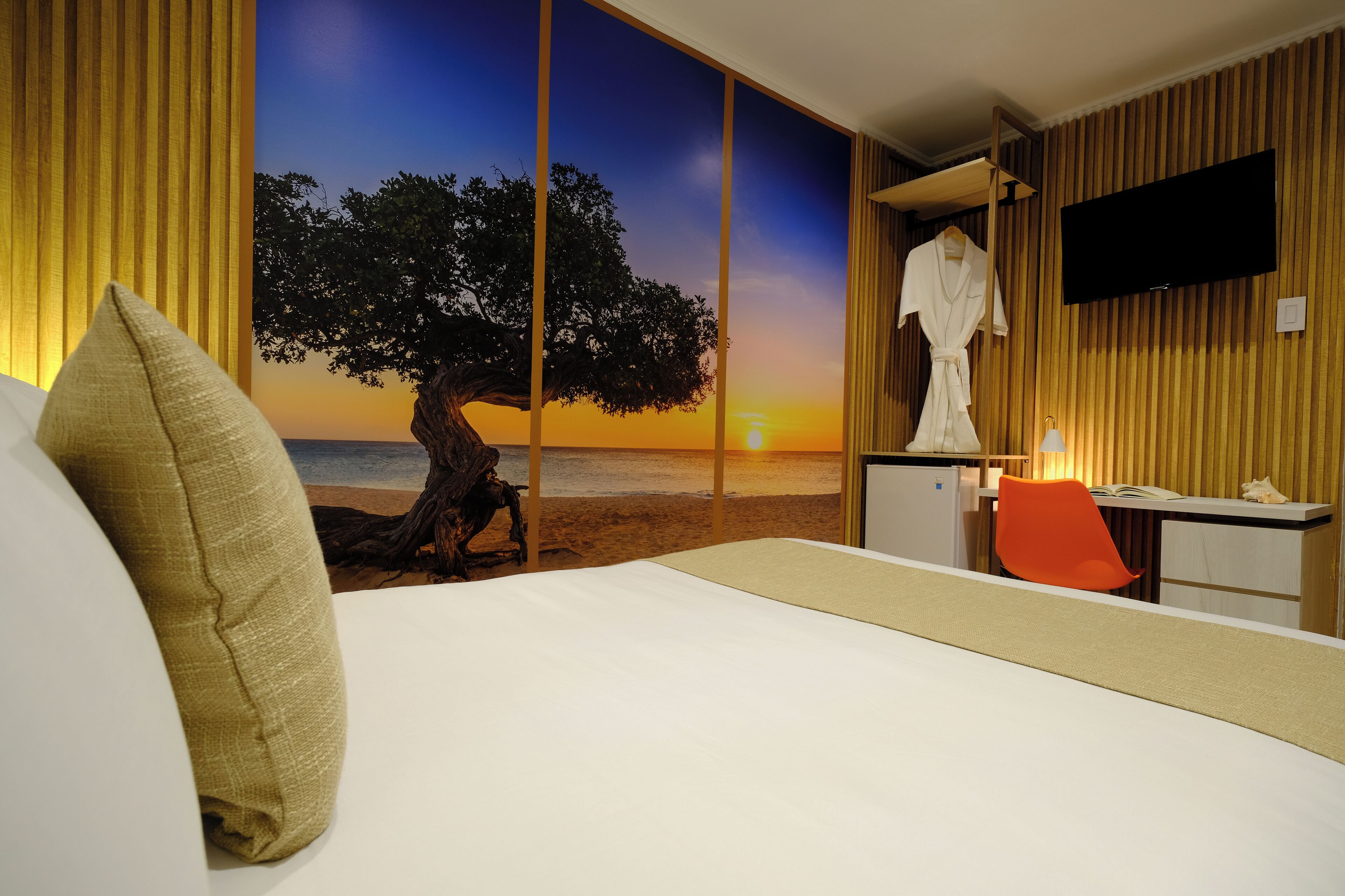 A hotel room with a bed, a desk, a TV, and a view of a sunset over the ocean.