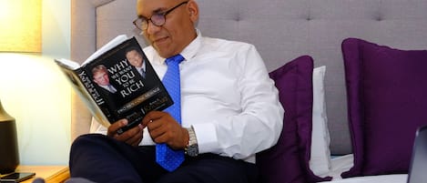 A man reading a book on a bed with a laptop and a smartphone on the bedside table.