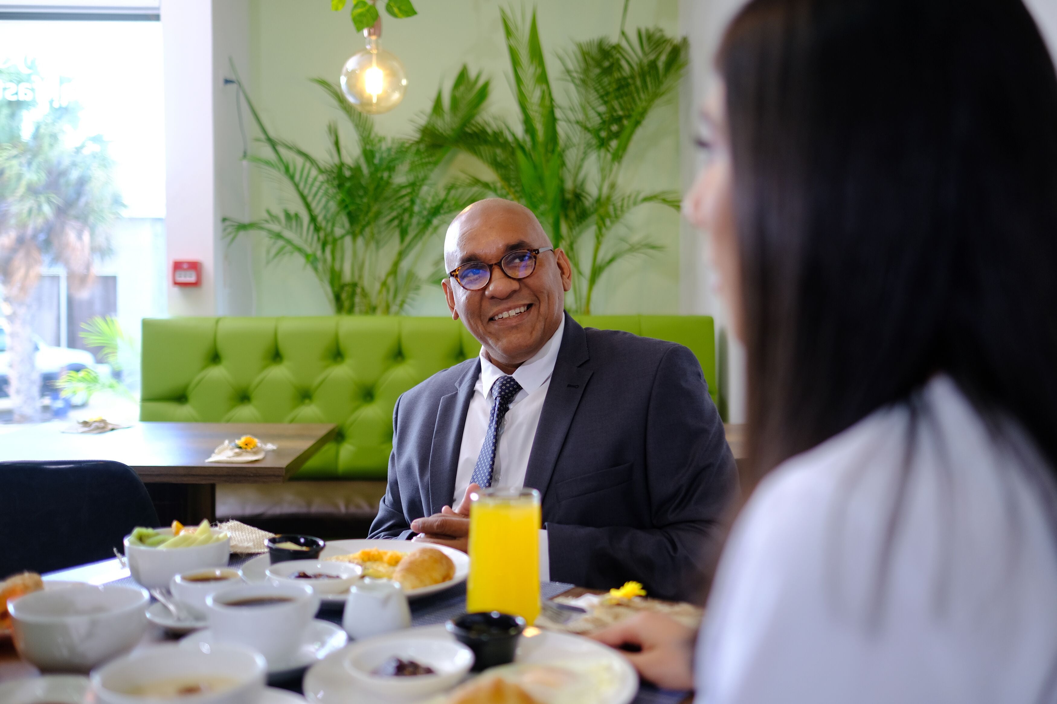 Un comedor con un banco acolchado verde, una mesa dispuesta con platos de comida y un vaso de zumo de naranja.