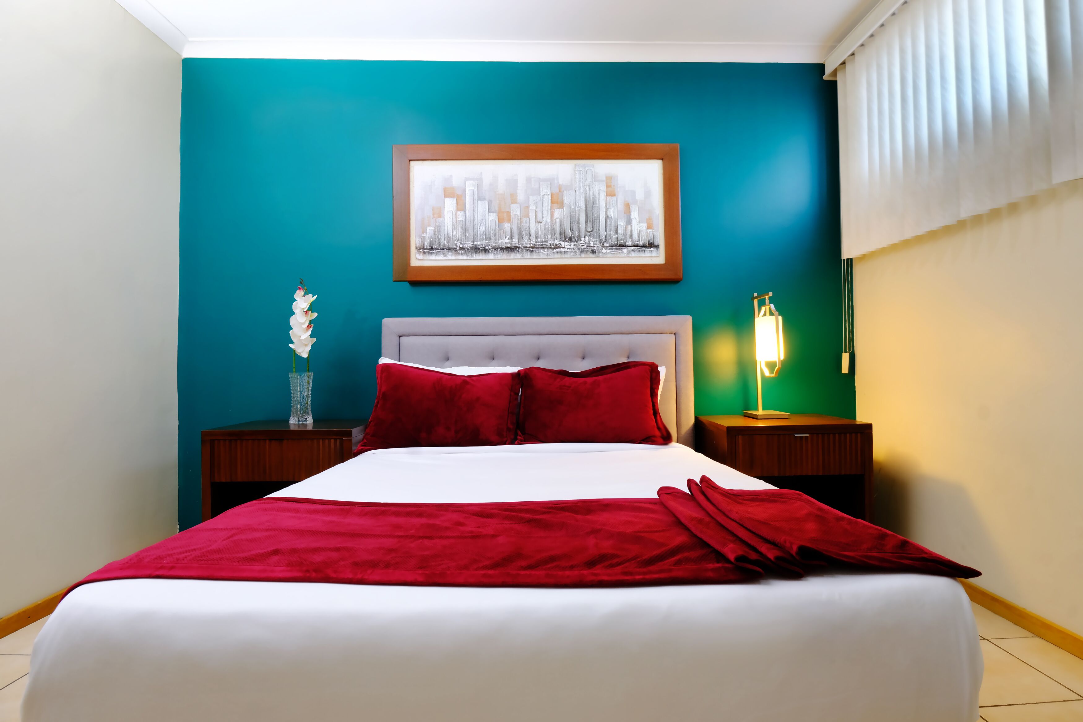 Ein Hotelzimmer mit einer türkisfarbenen Akzentwand, einem gerahmten Stadtbild und einem weißen Bett mit roter Bettwäsche. Ein Nachttisch mit einer Lampe steht daneben.