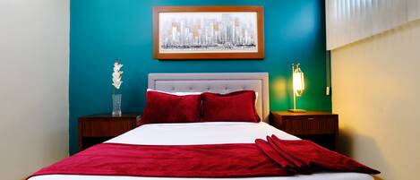 Ein Hotelzimmer mit einer türkisfarbenen Akzentwand, einem gerahmten Stadtbild und einem weißen Bett mit roter Bettwäsche. Ein Nachttisch mit einer Lampe steht daneben.