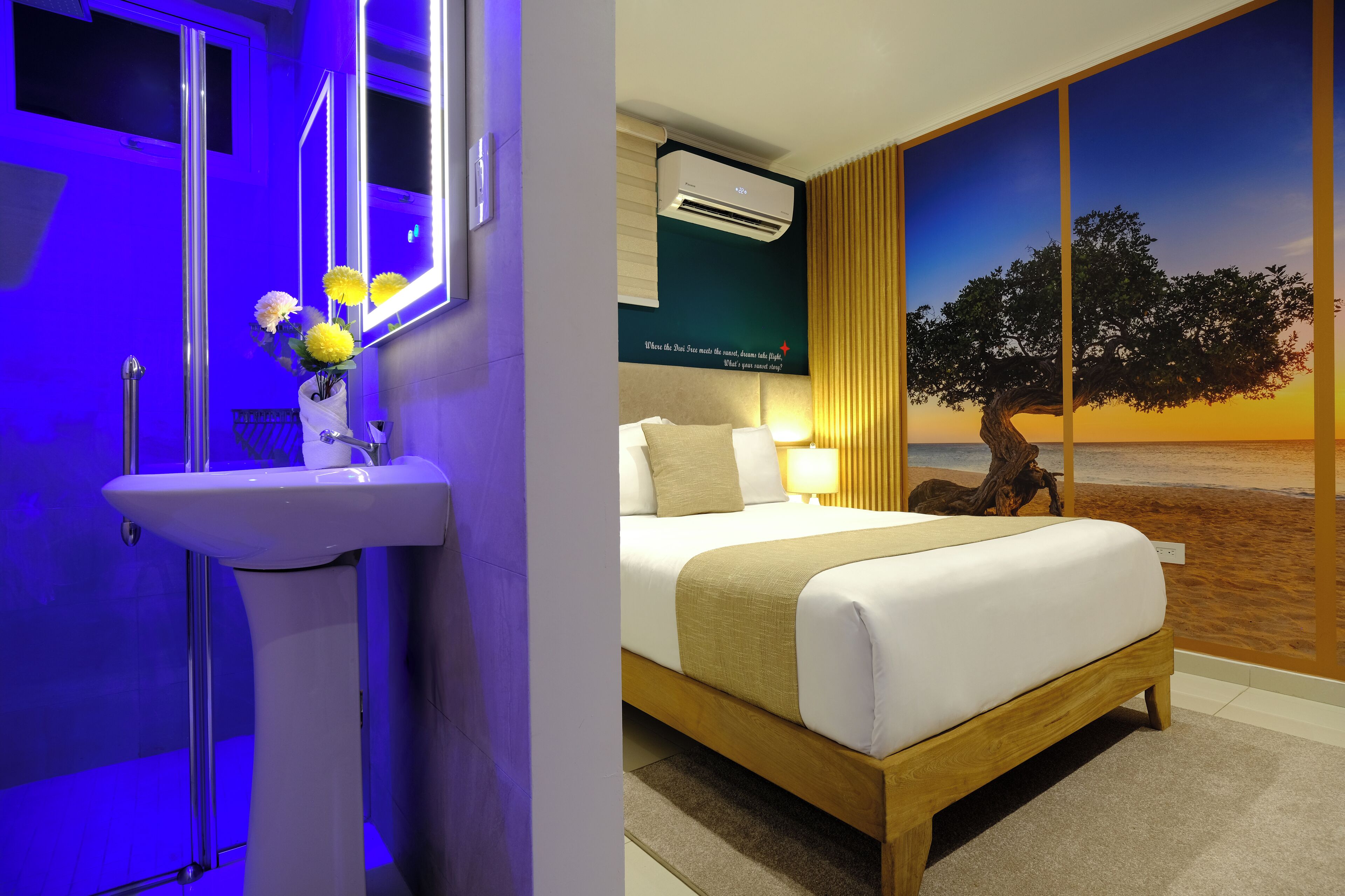 A hotel room with a bed, bedside table, lamp, and a large window with a view of a tree and sunset.