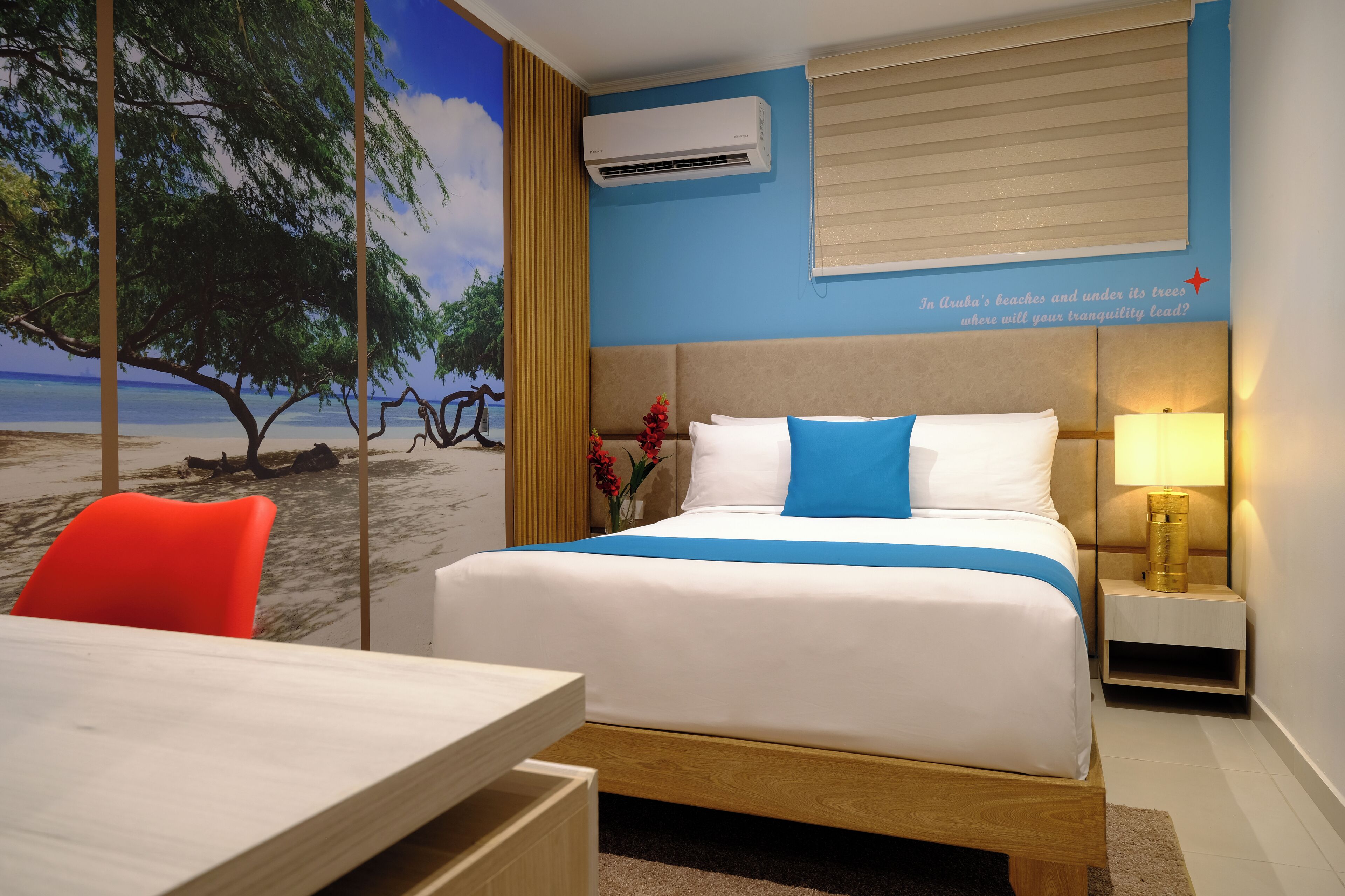 Quarto de hotel com cama, escrivaninha e cadeira. Um mural na parede retrata uma cena de praia.