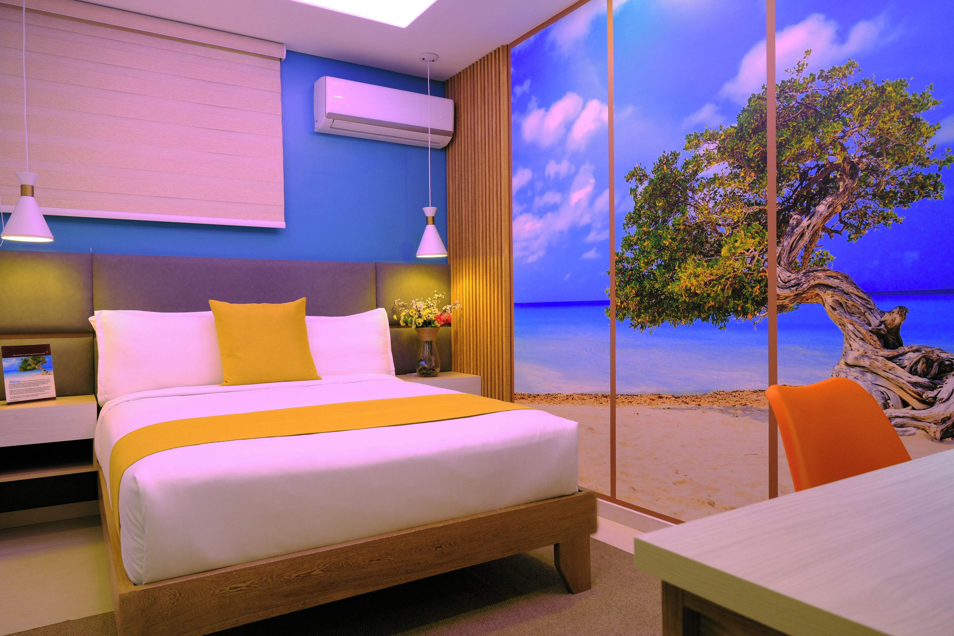 A modern bedroom with a large bed, a view of the beach through a window, and a table with an orange chair.