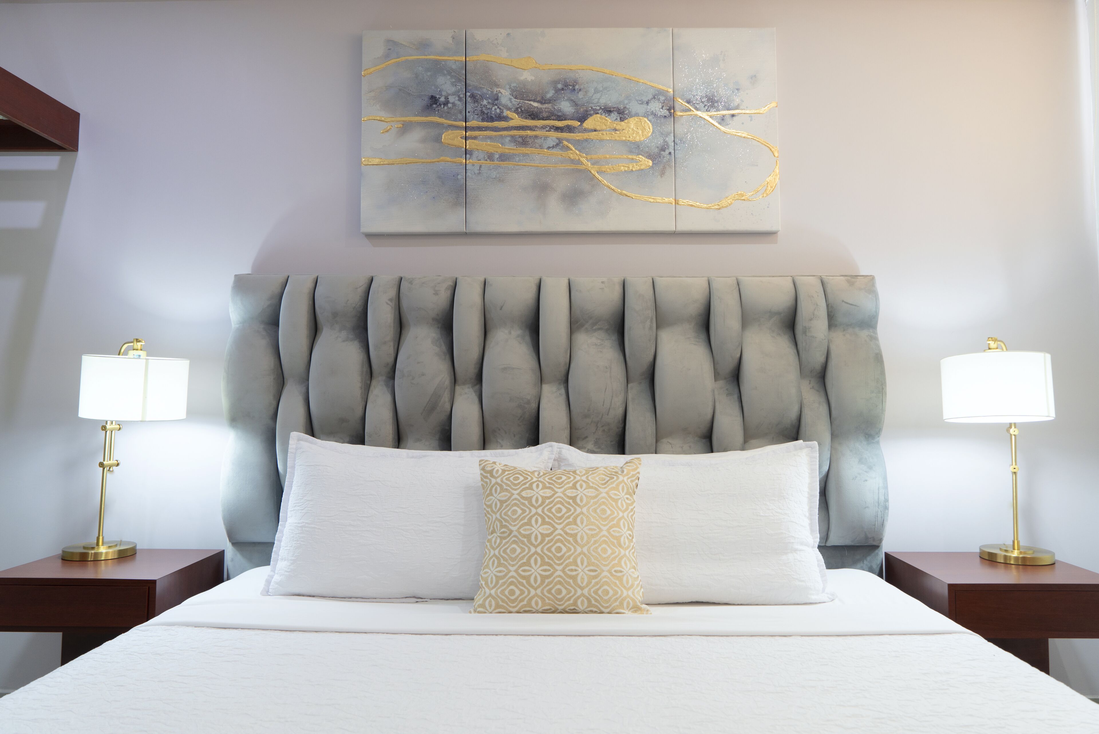 A hotel room with a tufted headboard, a large abstract painting, two bedside lamps, and a neatly made bed with white and patterned pillows.