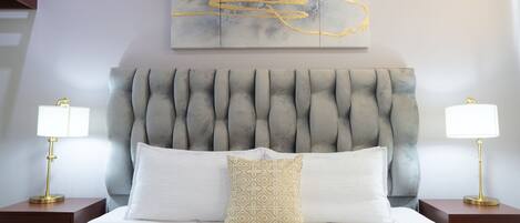 A hotel room with a tufted headboard, a large abstract painting, two bedside lamps, and a neatly made bed with white and patterned pillows.