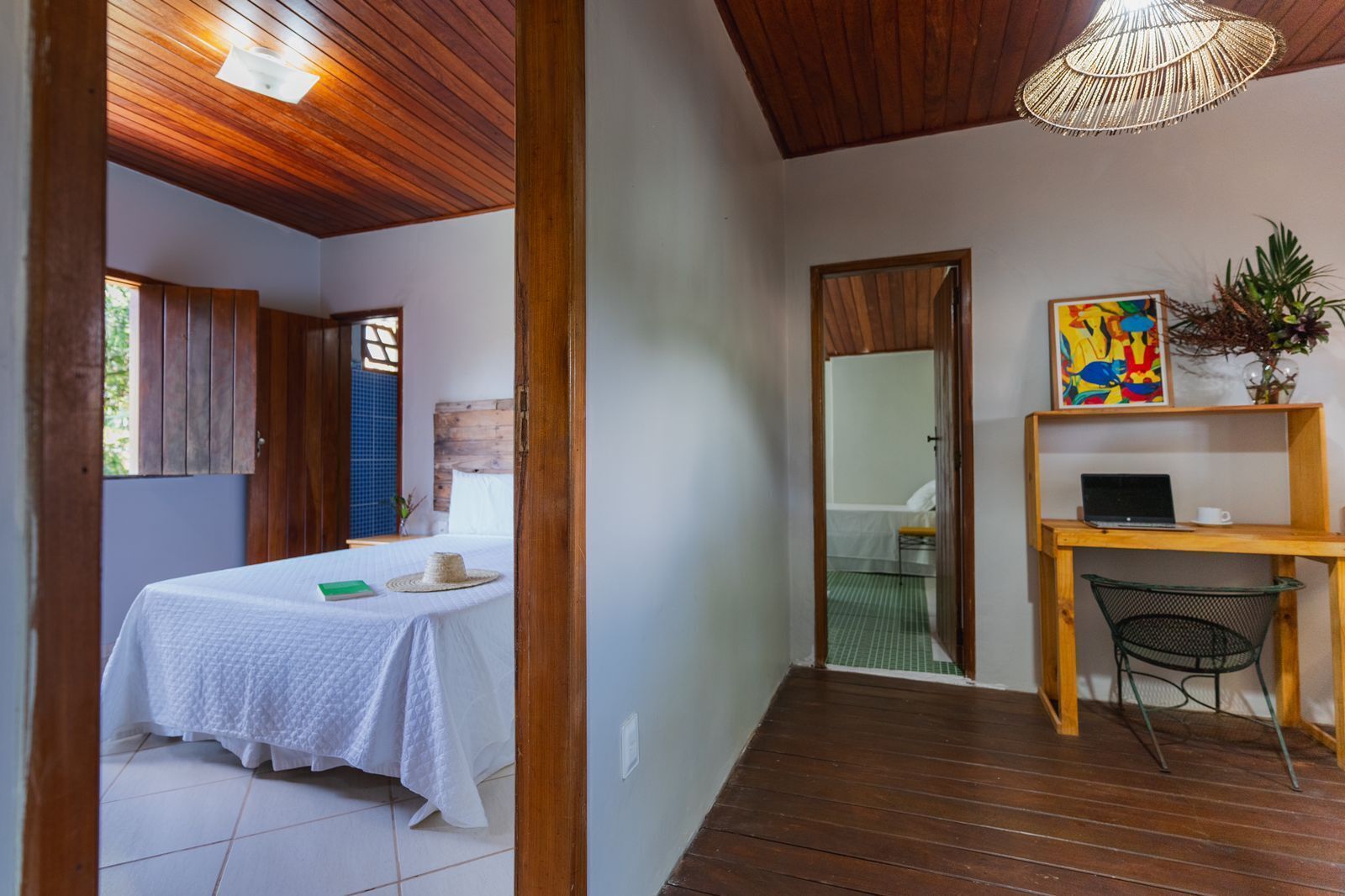 A room with a bed, a desk with a laptop, a chair, a plant, and a painting on the wall.