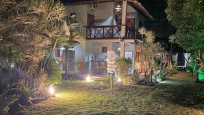 Edificio de dos pisos con balcón, rodeado de plantas tropicales e iluminado con luces artificiales.