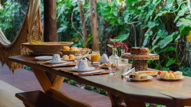 Mesa para comer al aire libre con alimentos para el desayuno, rodeada de abundante vegetación.