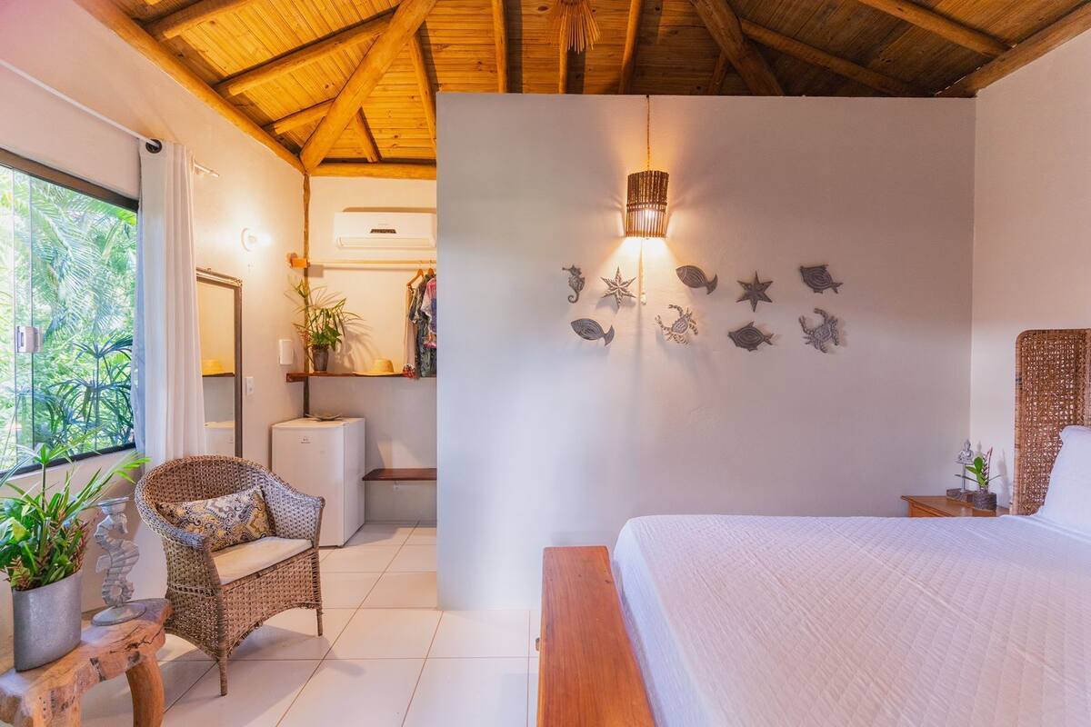 Une chambre à coucher comprenant un lit, une chaise en osier, une petite table, un luminaire fixé au mur et une plante en pot.