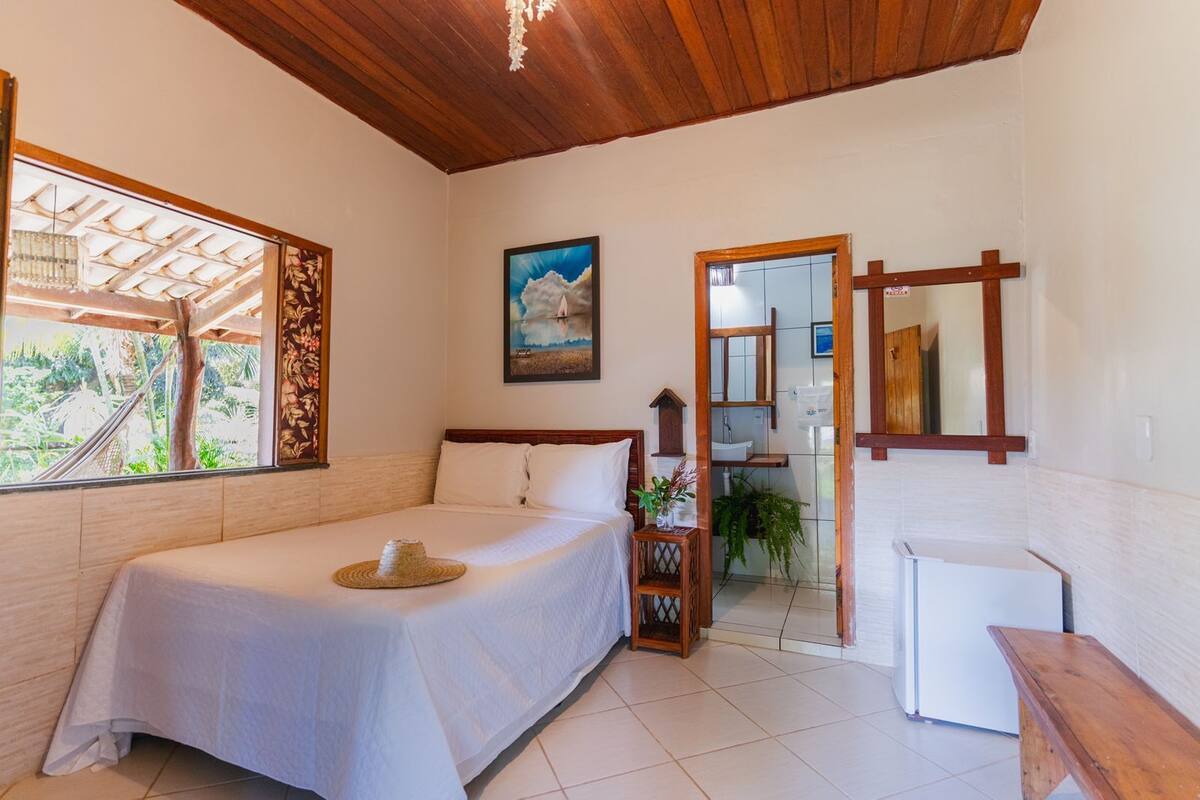 Une chambre à coucher avec un lit, une tête de lit en bois, une fenêtre donnant sur un paysage verdoyant, un tableau accroché au mur et une petite table sur laquelle est posée une plante.