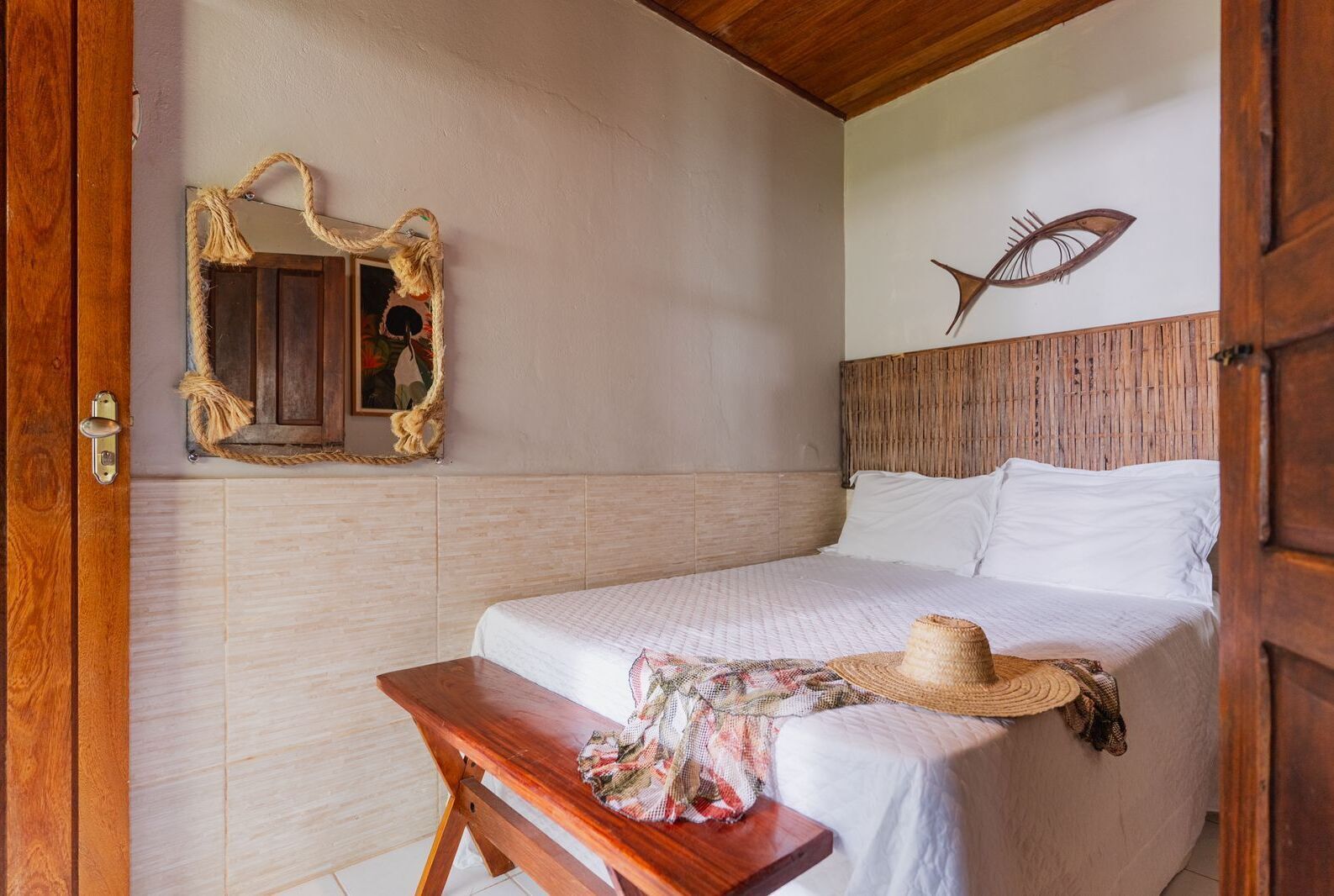 A bedroom with a wooden headboard, a bed with white linens, a wooden bench, a woven hat, and a mirror with a rope frame.