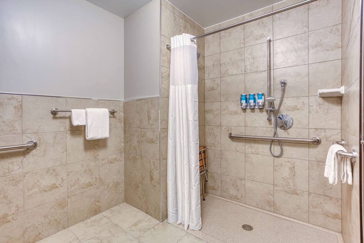 A shower area with beige tiles, a white shower curtain, and a handrail.