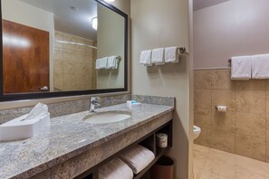 A bathroom with a granite countertop sink, a mirror, and a toilet. There are towels hanging on the wall and a trash can. - Drury Inn & Suites Cincinnati Northeast Mason (Mason)