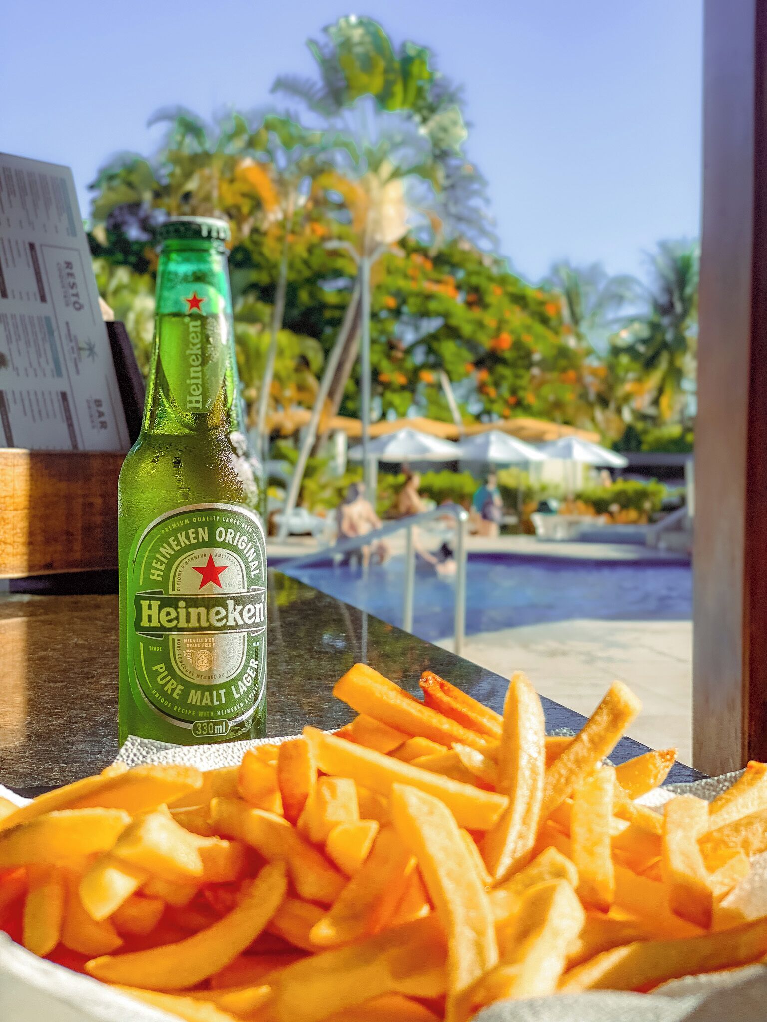 Una botella de cerveza Heineken y un bol de papas fritas sobre una mesa, con vista a una piscina y palmeras.
