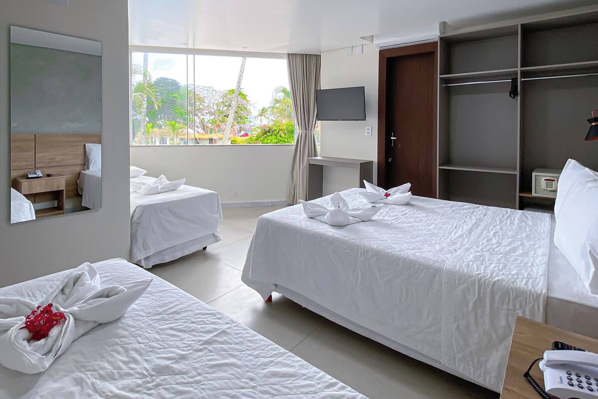 A hotel room with two beds, a TV, a wardrobe, and a balcony with a view of greenery.