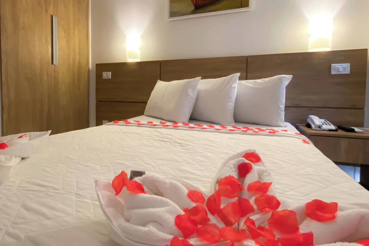 A neatly made bed with white linens and red rose petals, a wooden headboard, a painting of a boat on the wall, and a bedside phone.