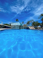 A swimming pool with clear blue water, surrounded by palm trees and a stone building.