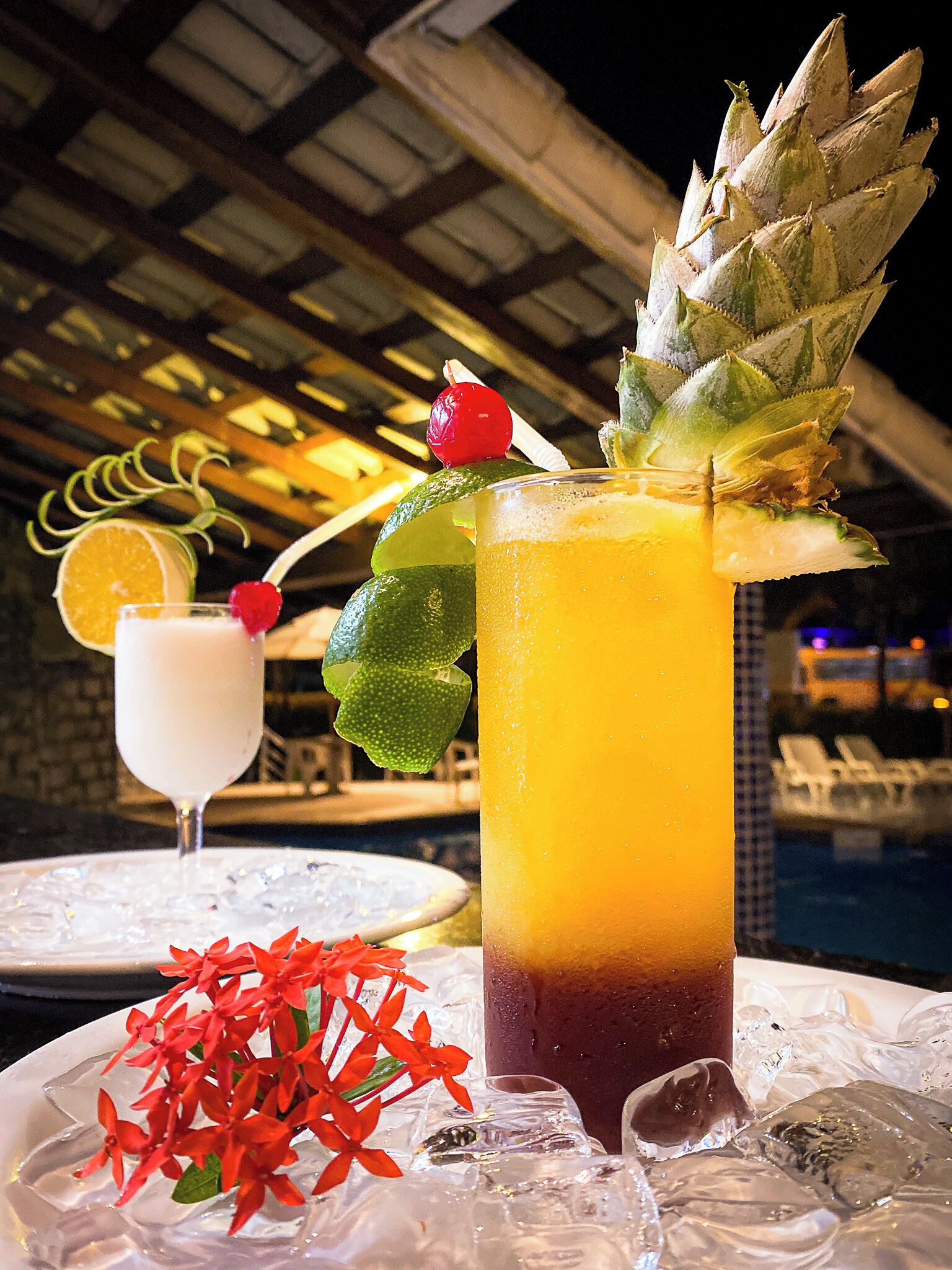 Um cocktail com um pedaço de abacaxi, uma fatia de limão e uma cereja numa taça, colocado sobre uma mesa com cubos de gelo e flores vermelhas.