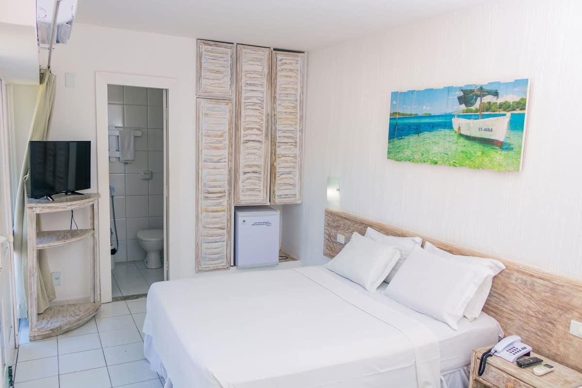 A hotel room with a bed, a TV, a small refrigerator, a painting of a boat on the wall, and a bathroom visible through an open door.