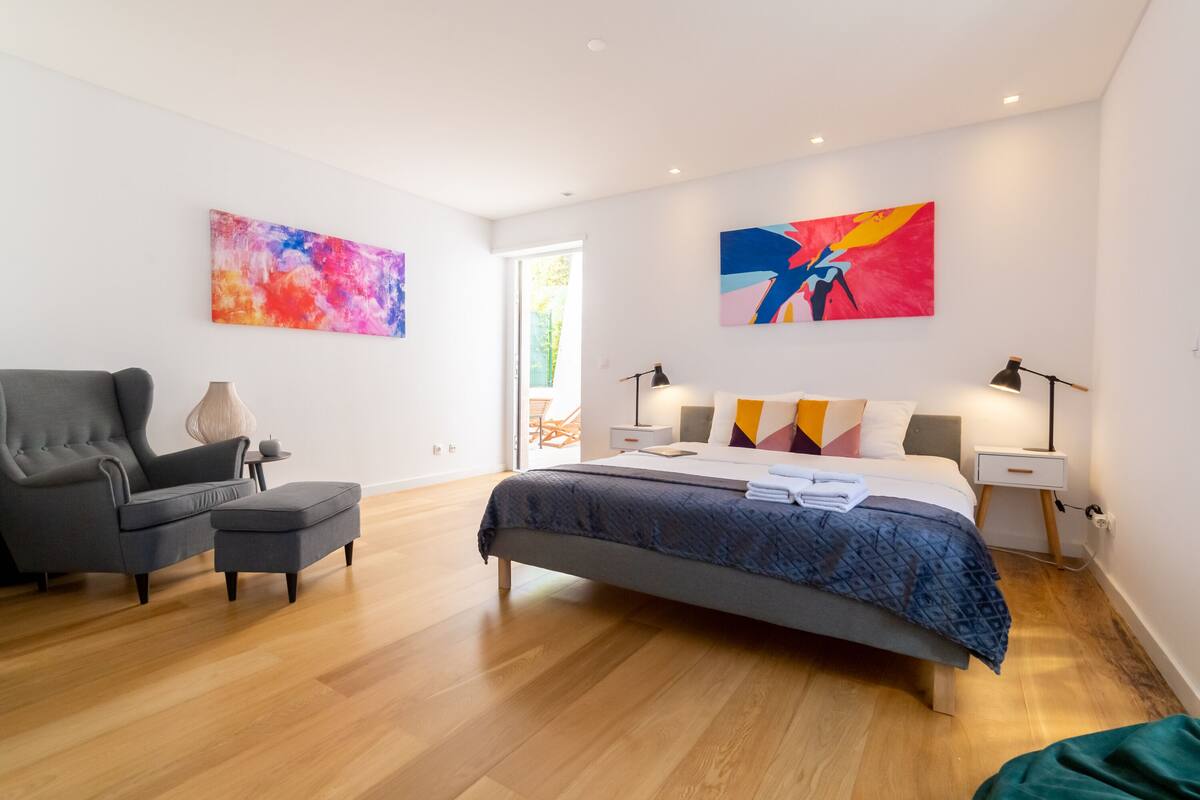 A modern bedroom with a bed, a grey armchair, a side table, and two abstract paintings on the walls.