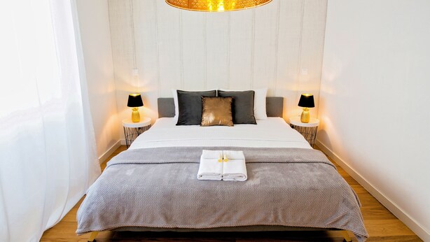 A neatly made bed with a grey bedspread and two pillows, a white towel folded in the center, and two bedside lamps on either side.