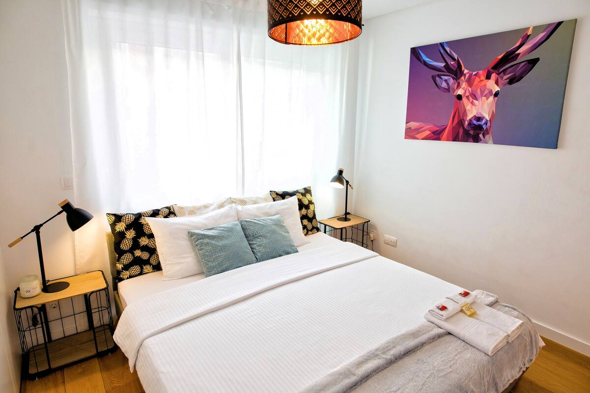 A neatly made bed with white bedding, a lamp on a bedside table, and a painting of a deer on the wall.