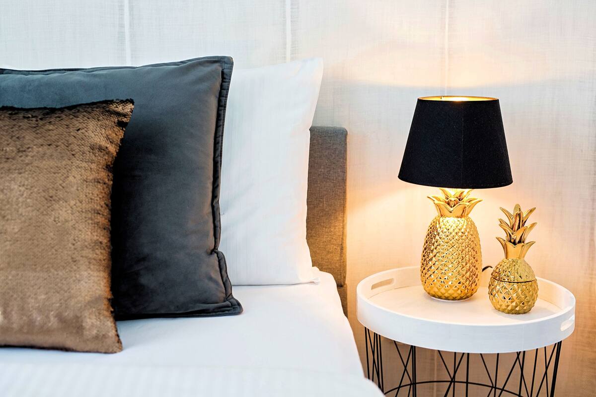 A neatly made bed with two pillows, a white wall, and a small round table with a lamp and pineapple-shaped vases.