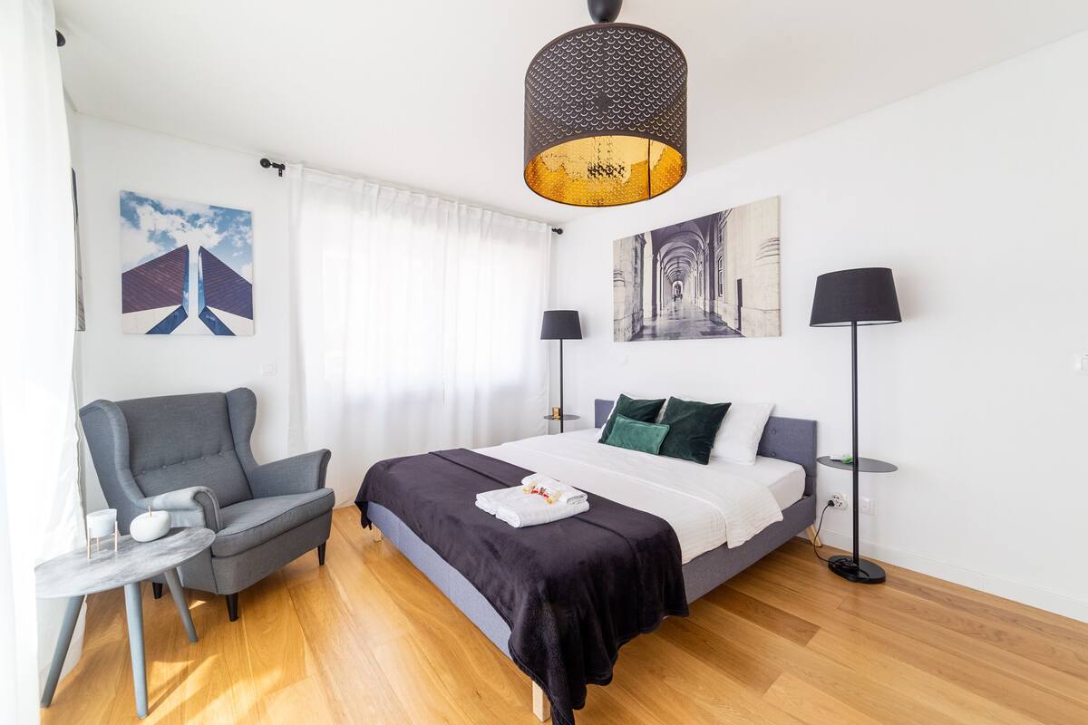 A modern bedroom with a bed, a chair, a small table, and artwork on the walls.