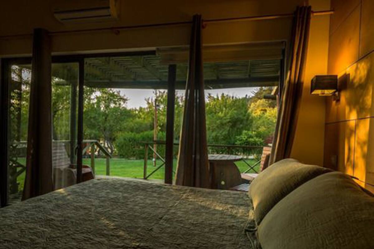 A bedroom with a large bed, a view of a garden, and a balcony with a table and chairs.
