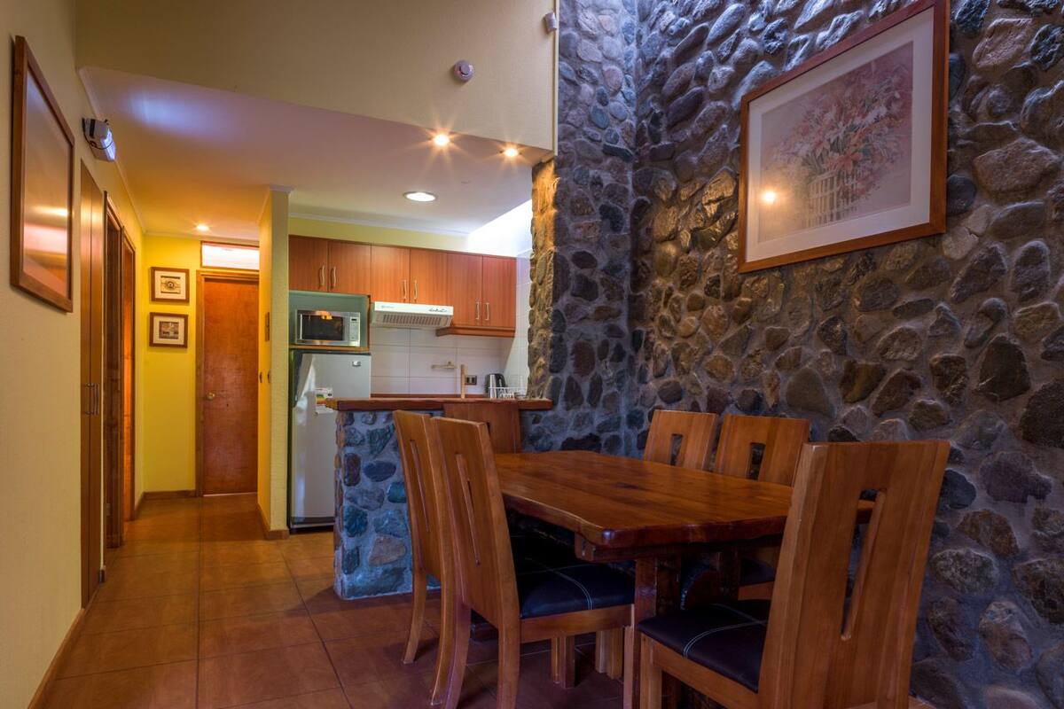 A dining area with a stone wall, wooden table, and chairs. A kitchen is visible in the background with wooden cabinets and a microwave.