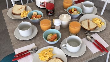 Desayuno con tostadas, huevos revueltos y fruta sobre una mesa, junto con una cafetera y tazas.