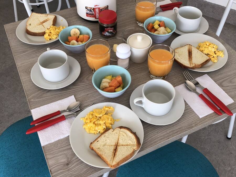 Desayuno con tostadas, huevos revueltos y fruta sobre una mesa, junto con una cafetera y tazas.