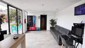 A modern hotel lobby with a reception desk, a seating area with a sofa and chairs, a TV mounted on the wall, and a view through large windows to the outside.