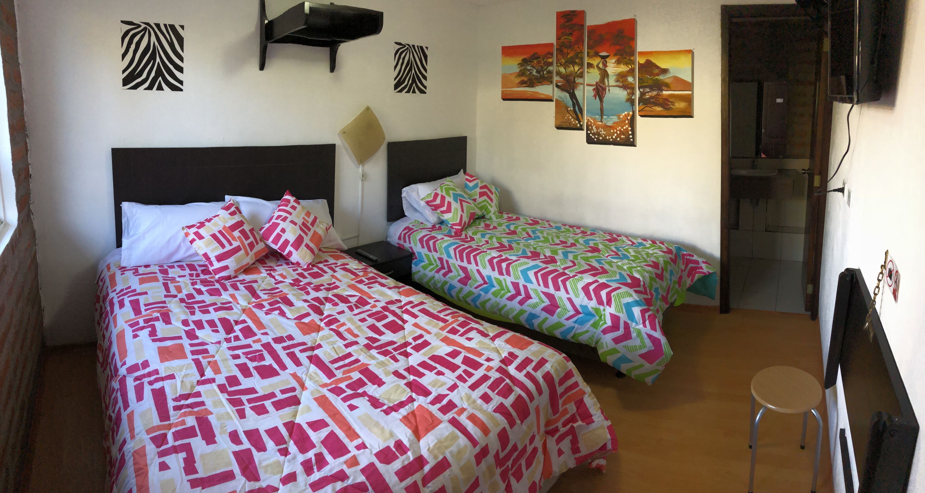 A room with two beds, each with patterned bedding, a small stool, and wall art.
