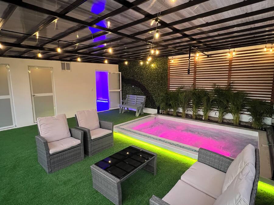 Un área de descanso al aire libre con césped artificial, mobiliario de mimbre y una piscina con iluminación rosa.