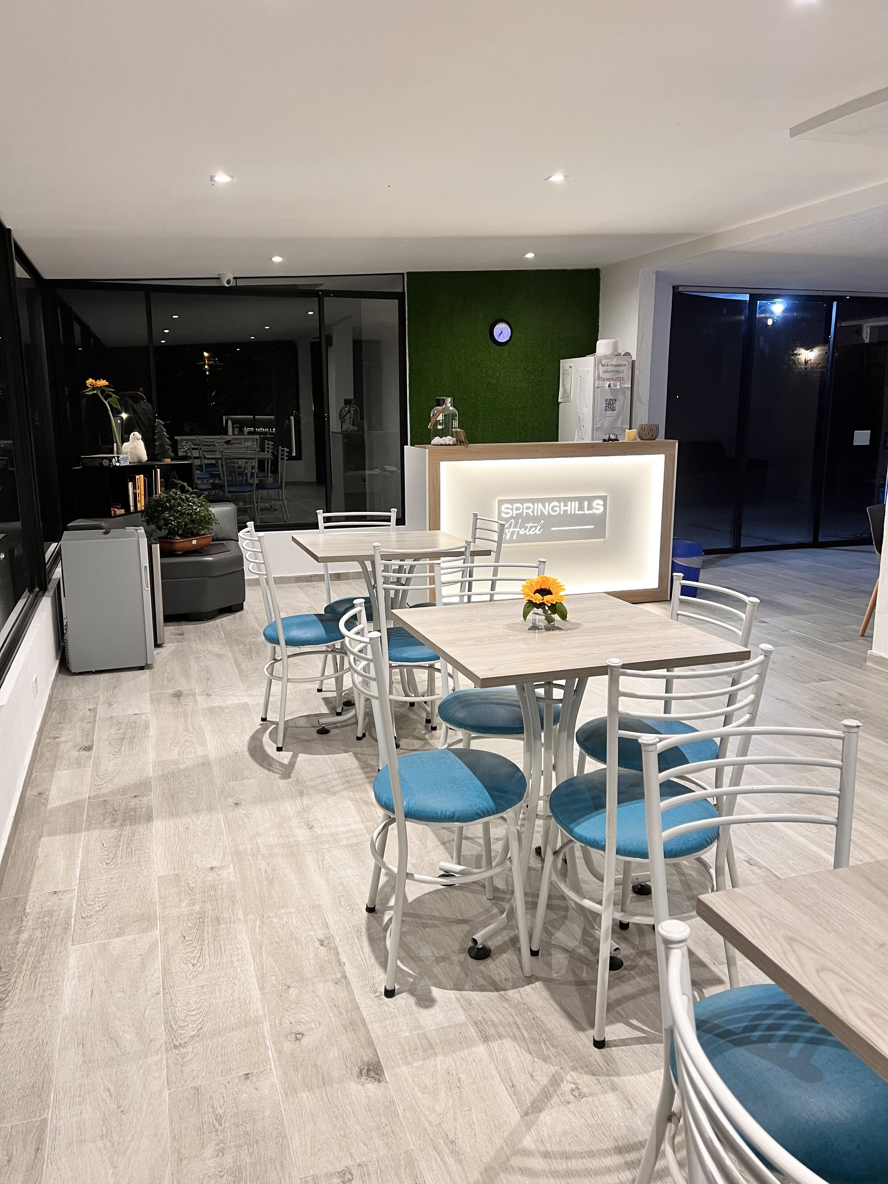A modern hotel lobby with a reception desk, seating area with blue cushioned chairs, and a green wall with a clock.