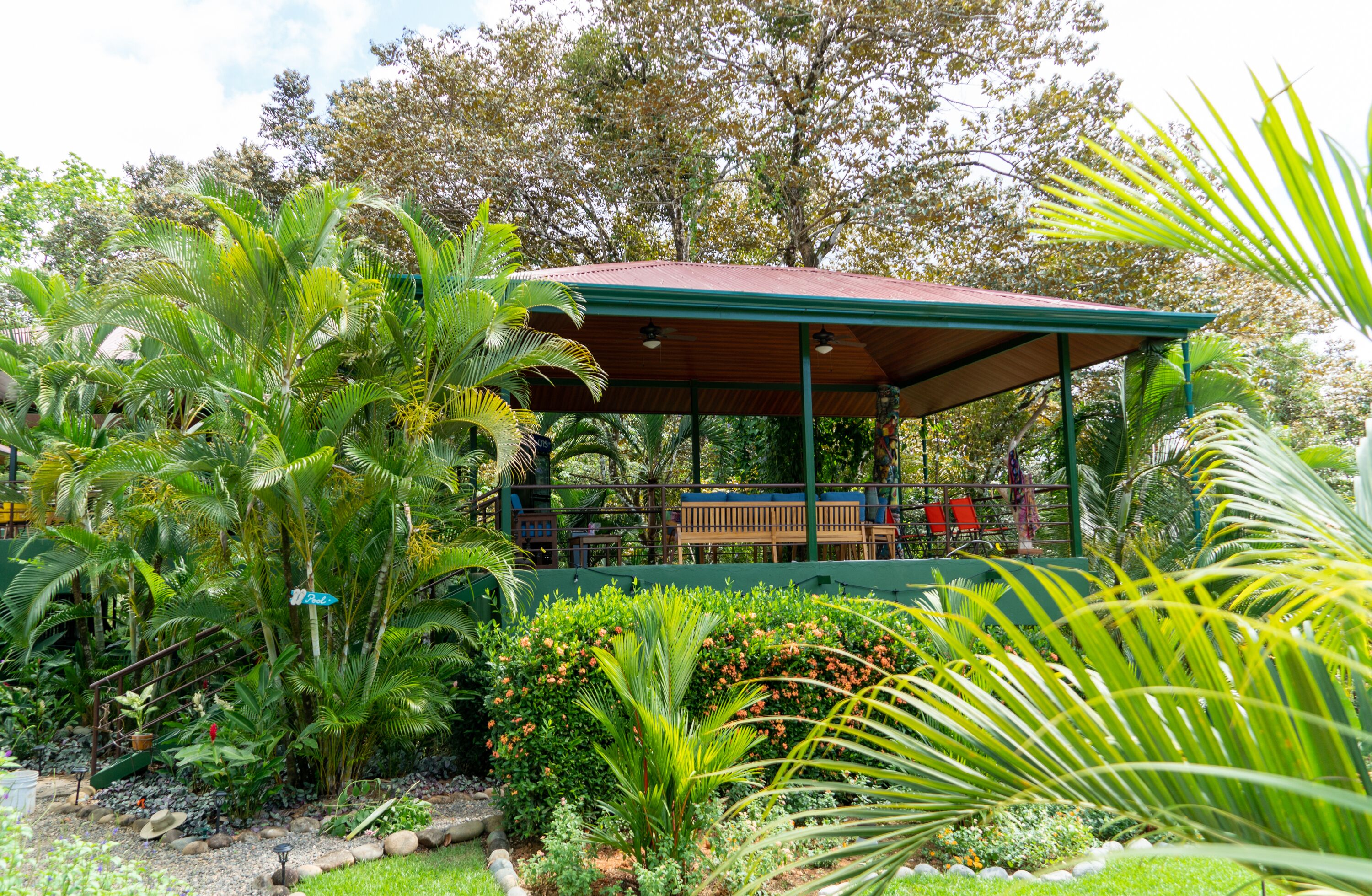 A covered outdoor seating area with benches and a wooden table, surrounded by lush tropical plants and trees.