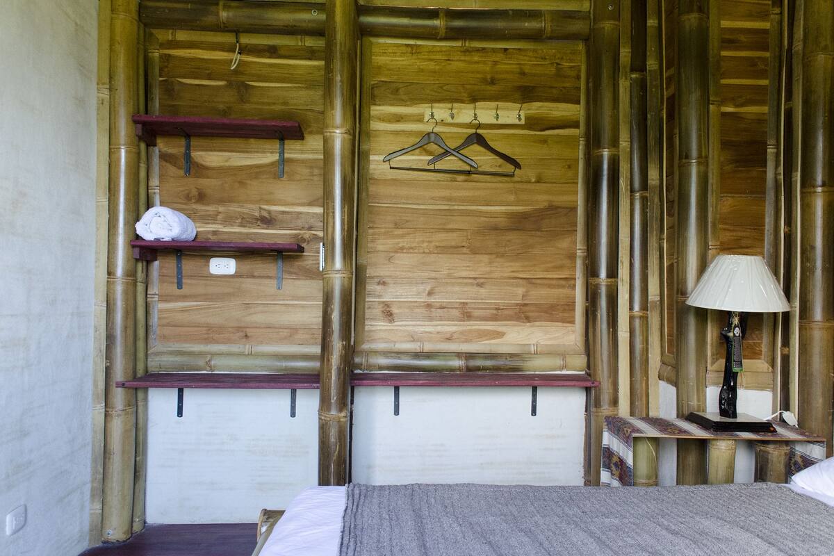 A wooden room with a bed, a nightstand, a lamp, a shelf with a towel, and two hangers on the wall.