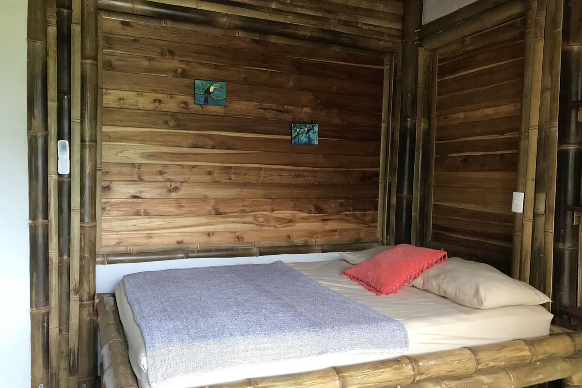 A bed with a grey blanket and red pillow in a wooden room with bamboo walls and a small painting.