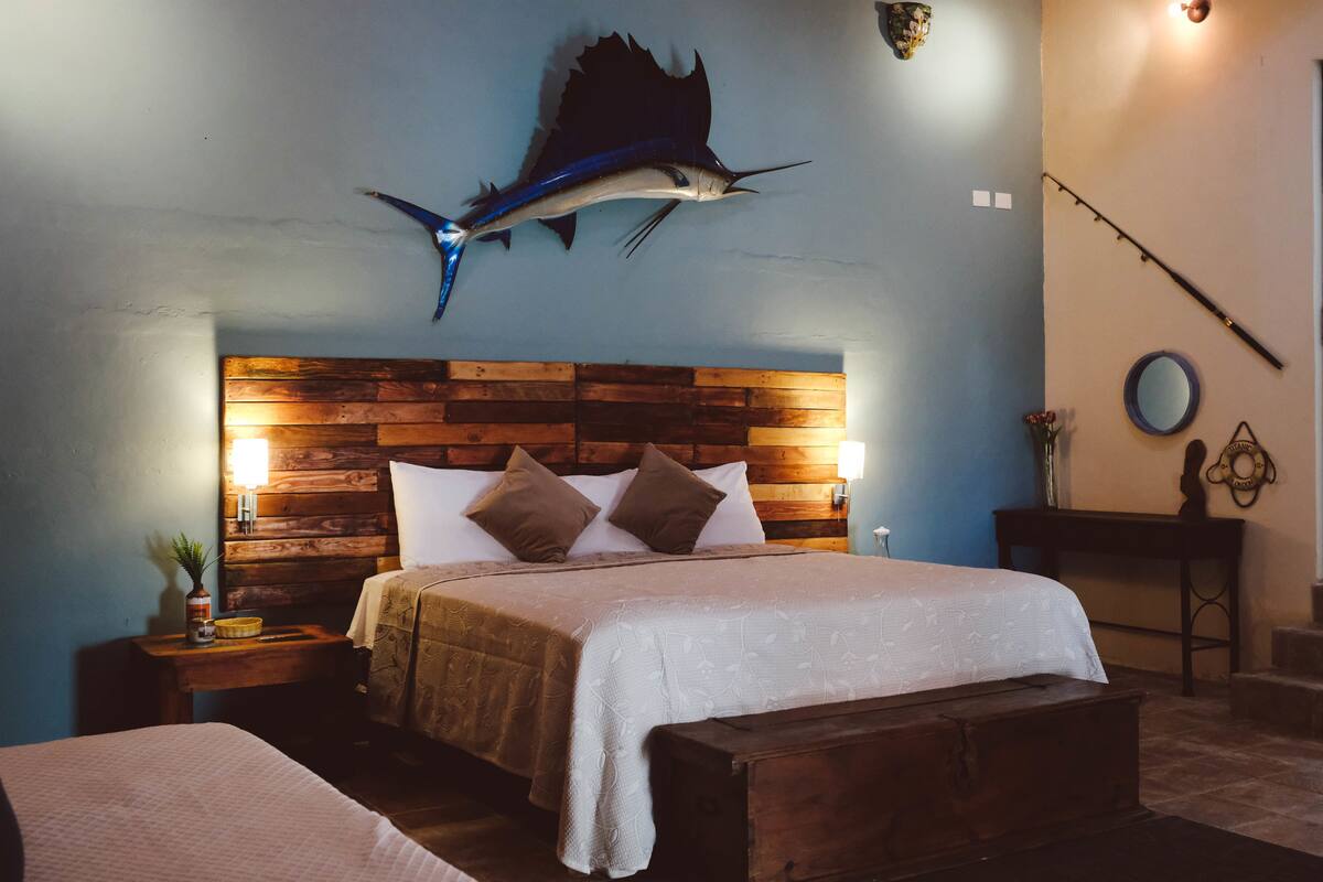 Une chambre à coucher avec une tête de lit en bois, un lit recouvert de draps blancs, une table de chevet avec une lampe et une canne à pêche fixée au mur.