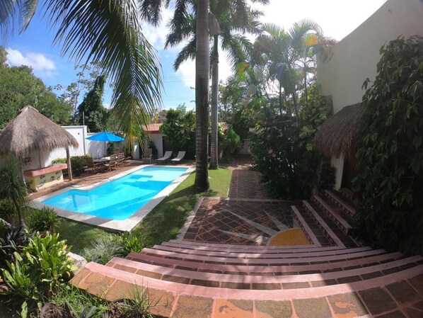 A swimming pool surrounded by a garden with palm trees and a thatched-roof structure.