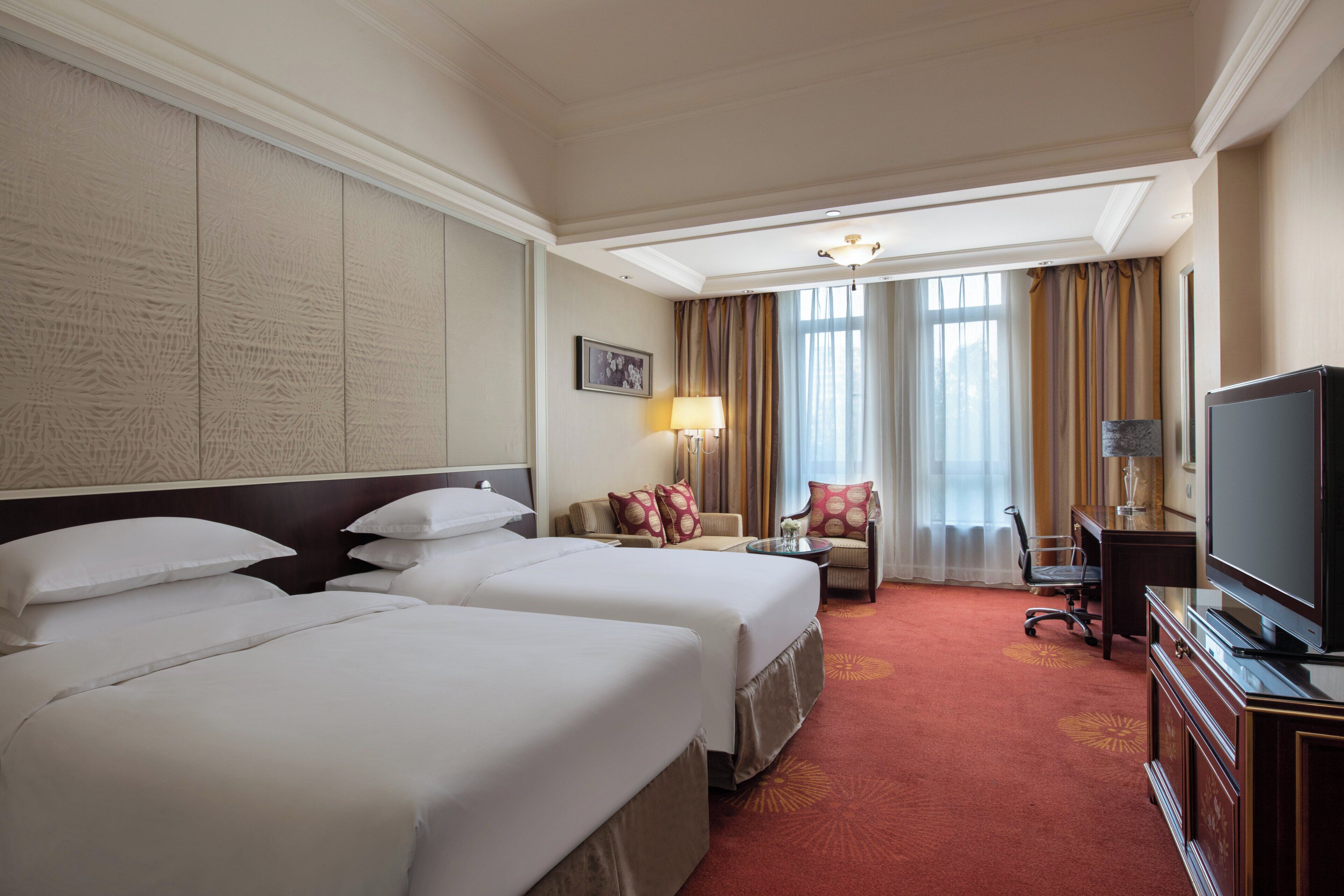 A hotel room with two beds, a desk with a television, a chair, and a lamp.