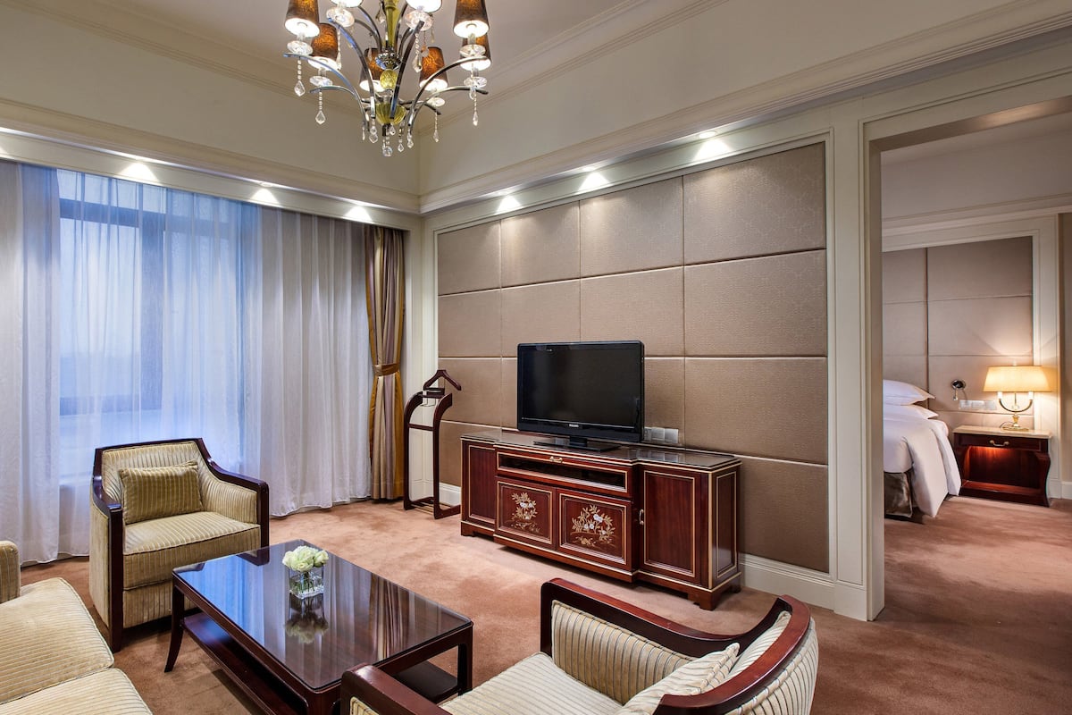 A hotel room with a TV, sofa, armchair, and a small table with a vase of flowers.