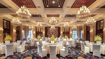 A banquet hall with multiple round tables, white chairs, chandeliers, and a patterned carpet.
