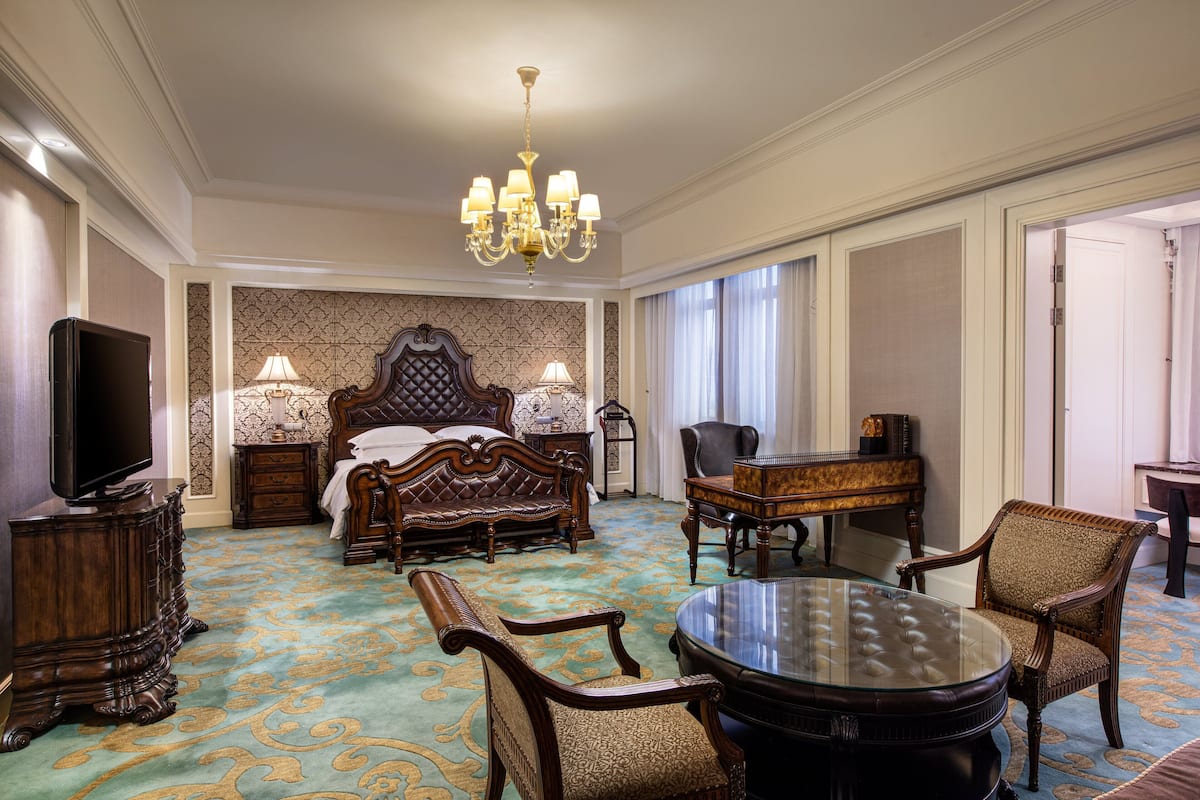 A spacious hotel room with a large bed, a sitting area with a piano, a television, and a chandelier.