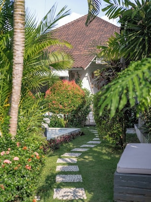 A garden pathway leading to a building with a tiled roof, surrounded by lush greenery and flowering plants. - Lila Boutik Residence (Canggu)