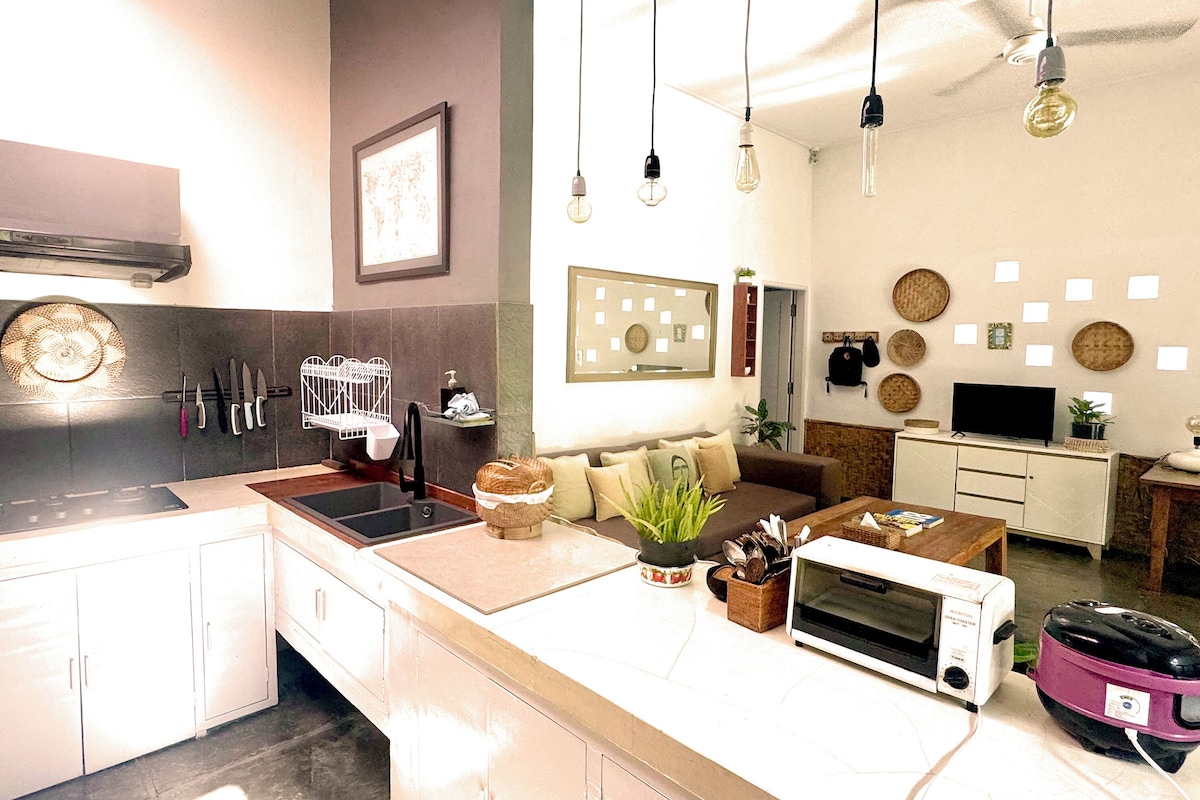 A modern kitchen with a built-in stove, sink, and microwave. There is a dining area with a wooden table and chairs. The kitchen features white cabinets and a black backsplash.