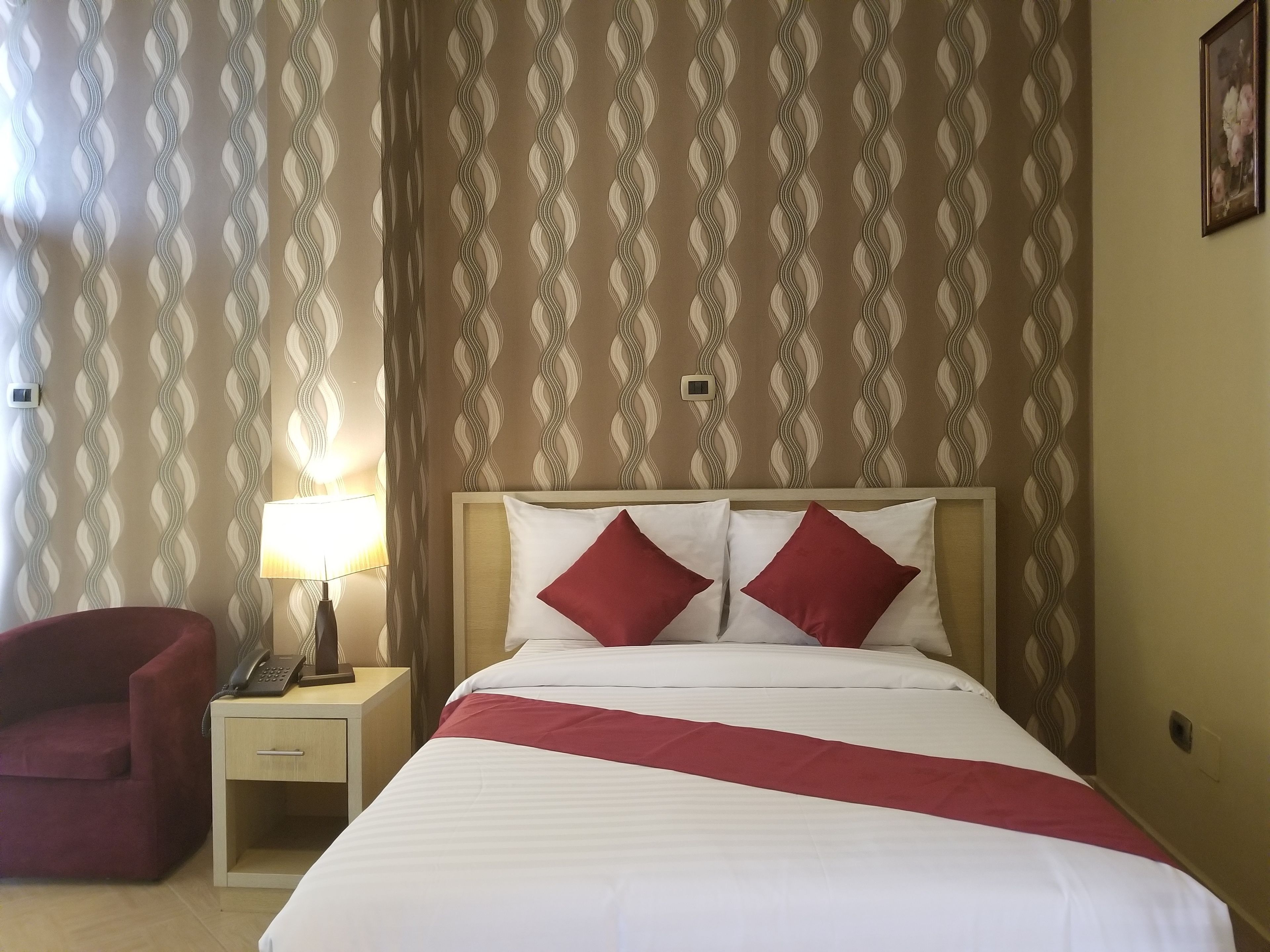 A hotel room with a bed, a nightstand, a lamp, a chair, and a wall with a wavy pattern.