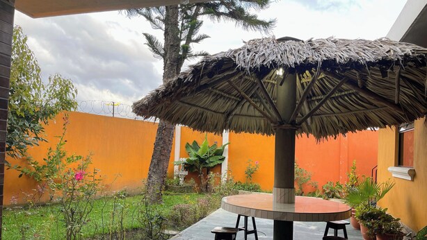 An outdoor patio with a round table, two stools, a thatched umbrella, potted plants, and a palm tree. - Casa hermosa a 20 minutos del aeropuerto (Guatemala City)